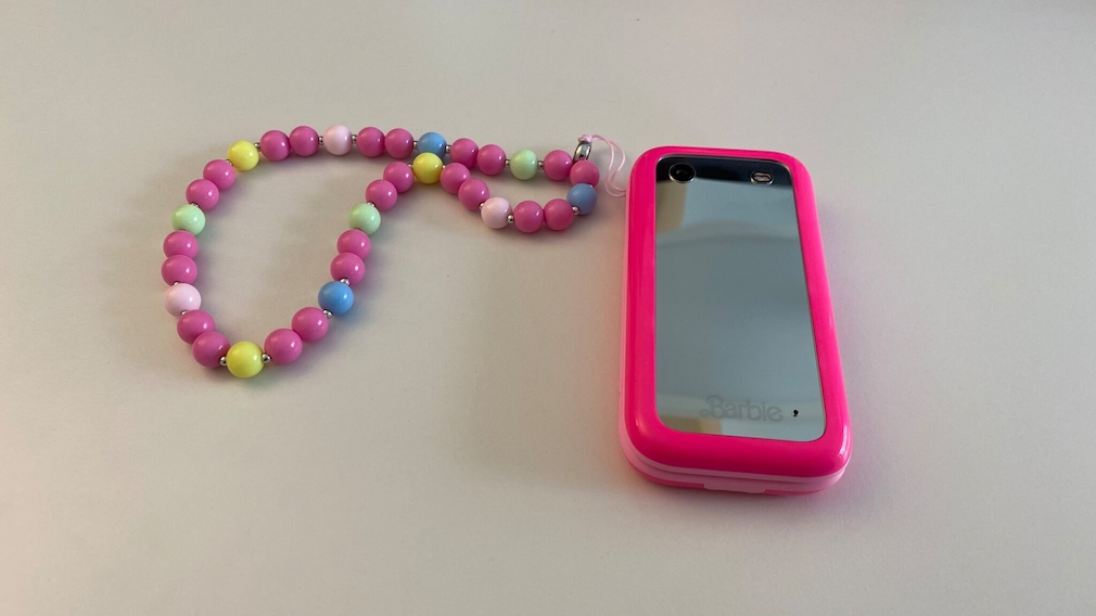 Barbie Phone liegend auf einem weißen Untergrund. Die Kette mit bunten Perlen ist an dem Handy befestigt. Das Handy liegt mit dem Spiegel nach oben.