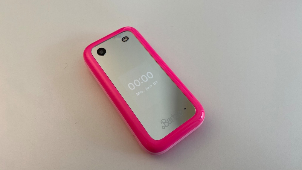 Barbie Phone zusammengeklappt auf einem weißen Untergrund liegend. Die Seote mit der Kamera, dem Blitz und dem Spiegel liegt oben. Auf dem Spiegel ist eine Uhrzeit und das Datum zu lesen.