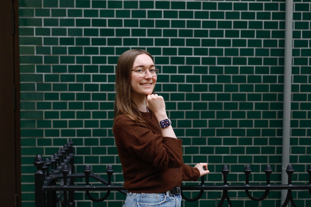 Frau mit der amazfit Active am Handgelenk vor einer Wand mit dunkelgrünen Backsteinen. Sie trägt einen braucnen Cardigan und eine hellblaue Jeans. Sie steht seitlich mit dem Gesicht zur Kamera. Sie lächelt.